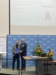 Zwei Männer auf einem Podium, wobei der Linke eine Urkunde in der Hand hält.